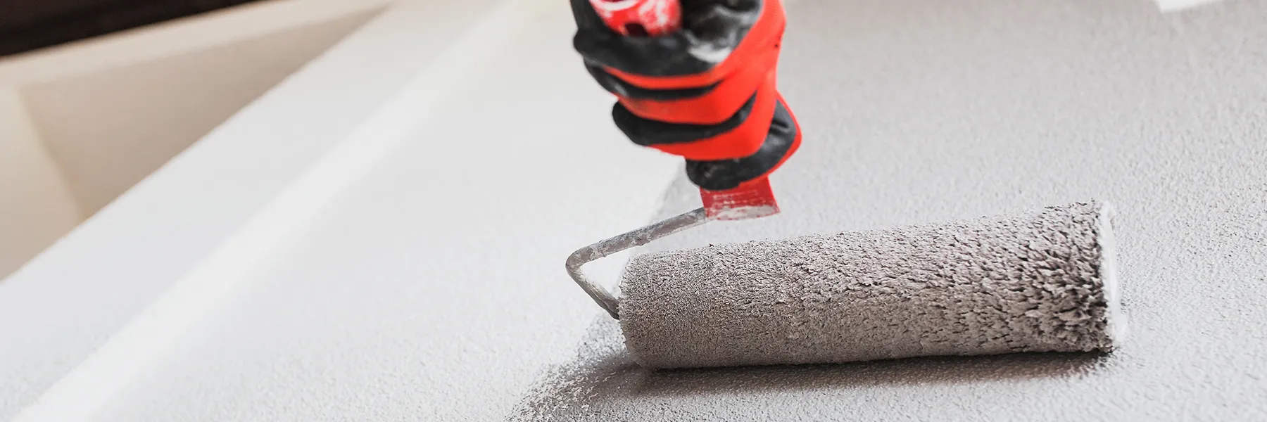 Close up of person wearing red glove applying grey paint with a roller to wall in commercial space.